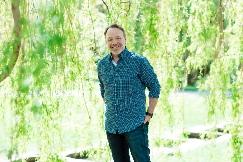 Thor Magne står oppstilt og smiler under et tre med hengende greiner. Solen skinner gjennom de lyse bladene.